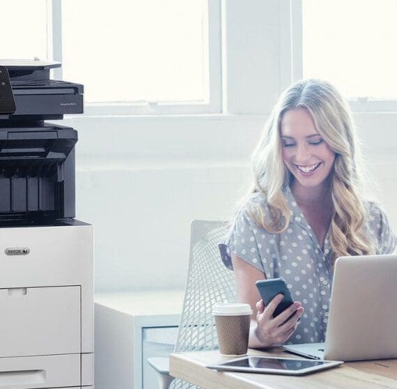 Una persona guarda felicemente il suo smartphone mentre è seduta alla scrivania con un computer portatile. Una stampante multifunzione Xerox VersaLink B605 è visibile accanto alla scrivania, in un ufficio ben illuminato.