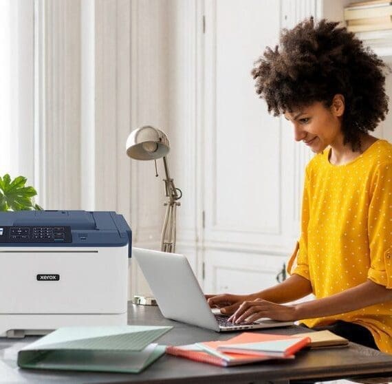 Una persona con i capelli ricci sorride mentre scrive su un computer portatile seduto alla scrivania, con una stampante compatta Xerox C310 posizionata accanto al computer portatile, a dimostrazione di una moderna configurazione di ufficio domestico per la produttività.