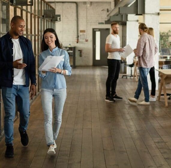 Due colleghi camminano lungo un corridoio in un moderno ufficio open space, tenendo in mano dei documenti e sorridendo, con altri colleghi visibili sullo sfondo, a rappresentare un ambiente di lavoro dinamico e collaborativo.