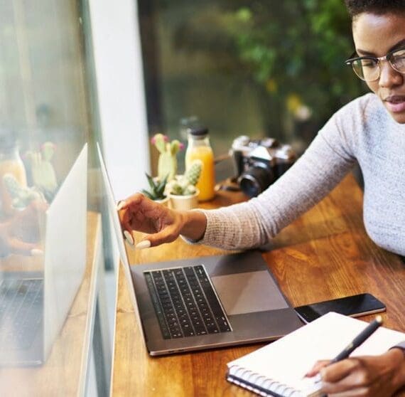 Una persona che indossa gli occhiali lavora a un tavolo di legno, guardando lo schermo di un laptop e scrivendo su un quaderno, con uno smartphone nelle vicinanze, suggerendo un ambiente di lavoro produttivo